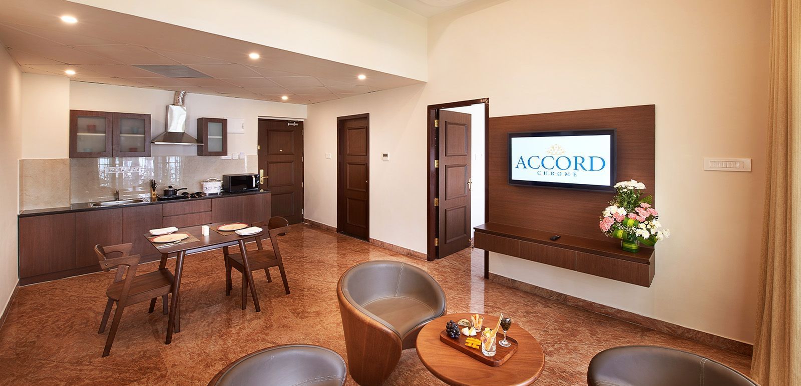 A wide angle shot of a living area in our suite featuring cosy seating arrangement, a wall mounted television and a kitchenette - Accord Chrome Airport Hotel